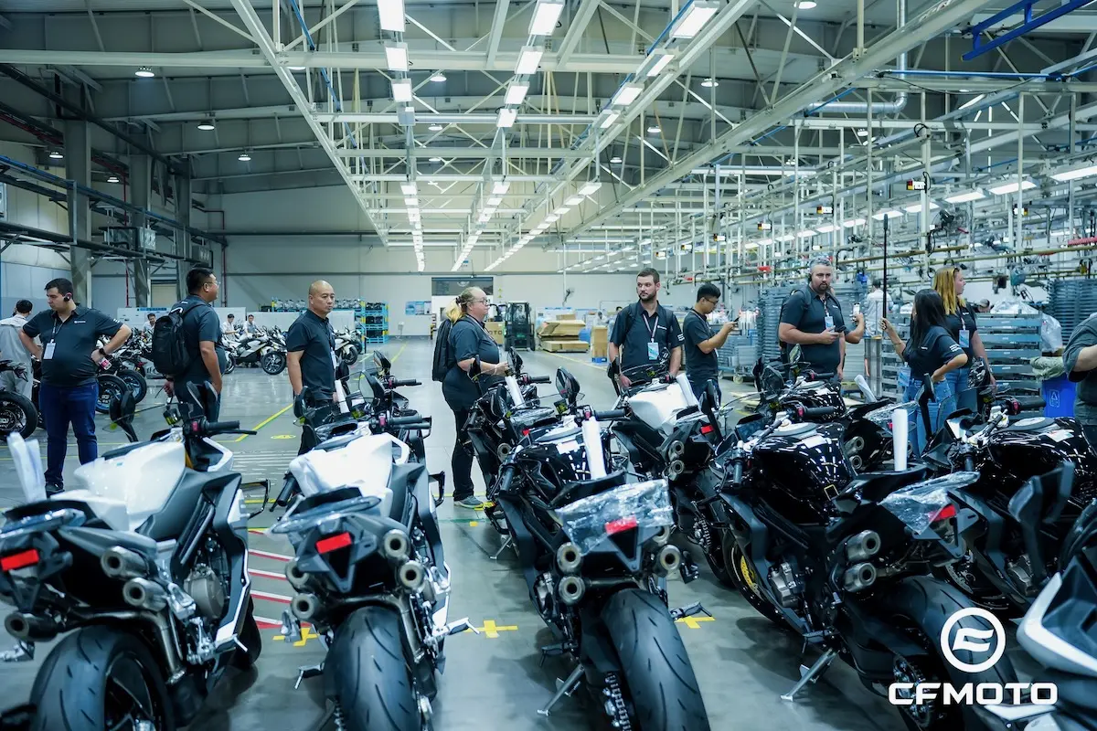 People stand and inspect rows of motorcycles inside a well-lit factory or assembly plant, with the CFMOTO logo visible in the lower right corner, highlighting the brand’s commitment to quality and ATV safety.