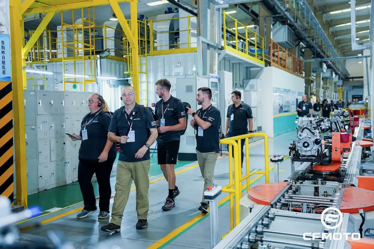 A group of people wearing CFMOTO shirts walk through a brightly lit manufacturing facility with machinery and equipment visible, emphasizing the brand's commitment to quality and ATV safety.