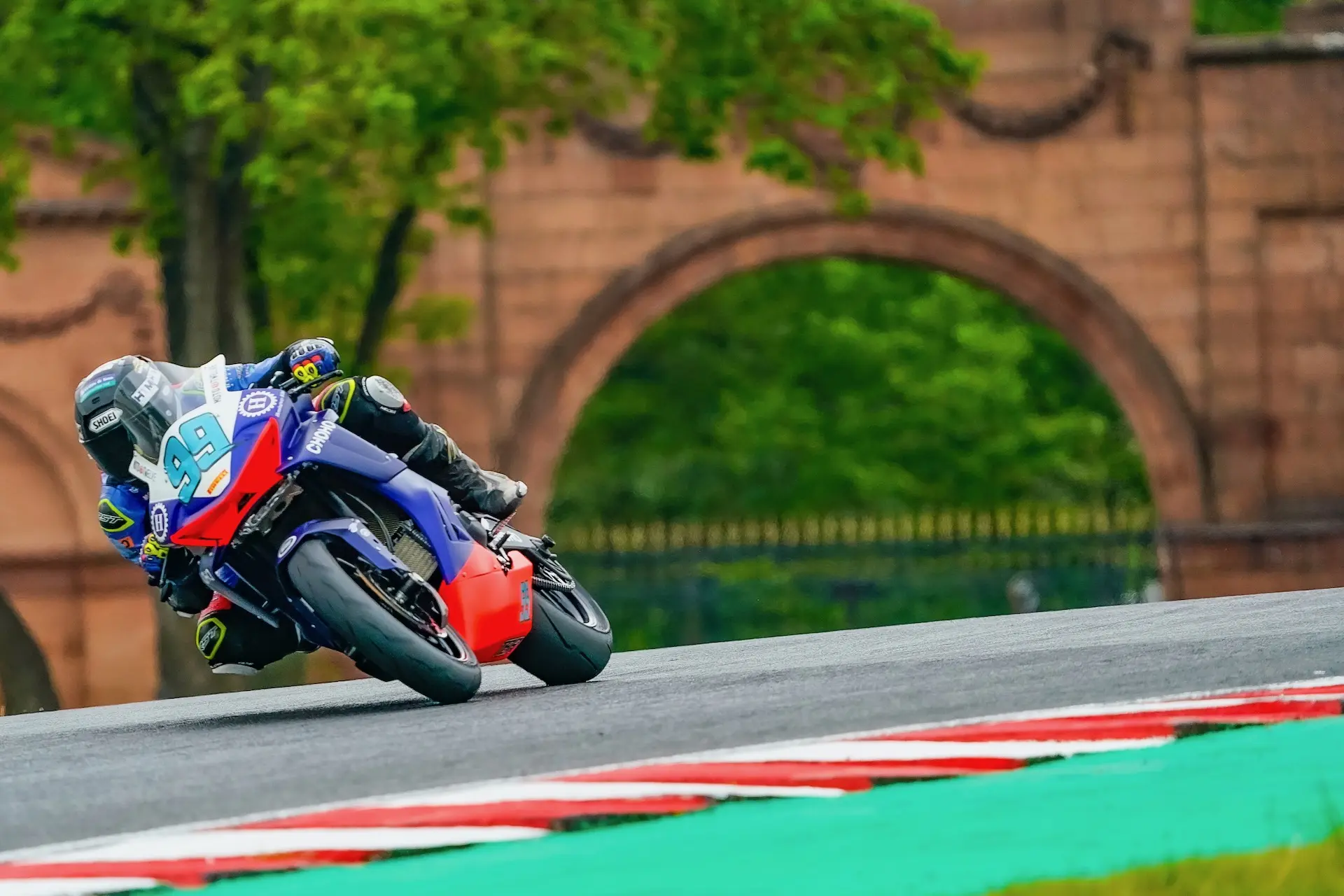 A motorcyclist in racing gear leans into a bend at Oulton Park race circuit, with green grass and a stone archway in the background.
