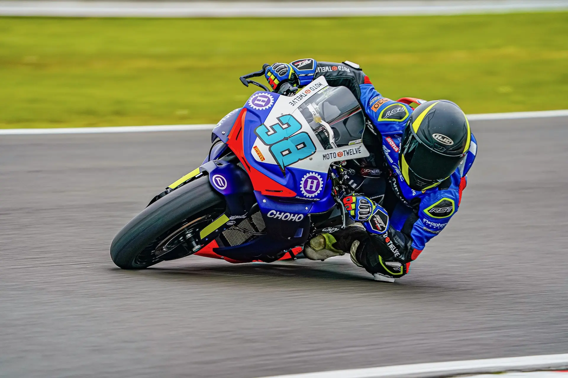 A motorcycle racer in blue and green kit, riding for CFMOTO, leans into a sharp bend at Oulton Park with the number 38 displayed on the bike.