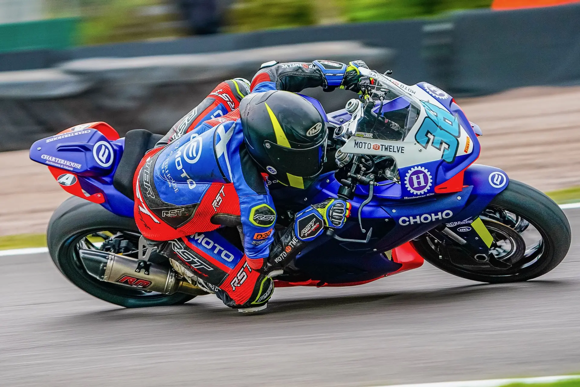 A motorcycle racer in blue and red gear leans sharply into a turn at Oulton Park racetrack, gripping the handlebars of a CFMOTO bike and wearing a black helmet.