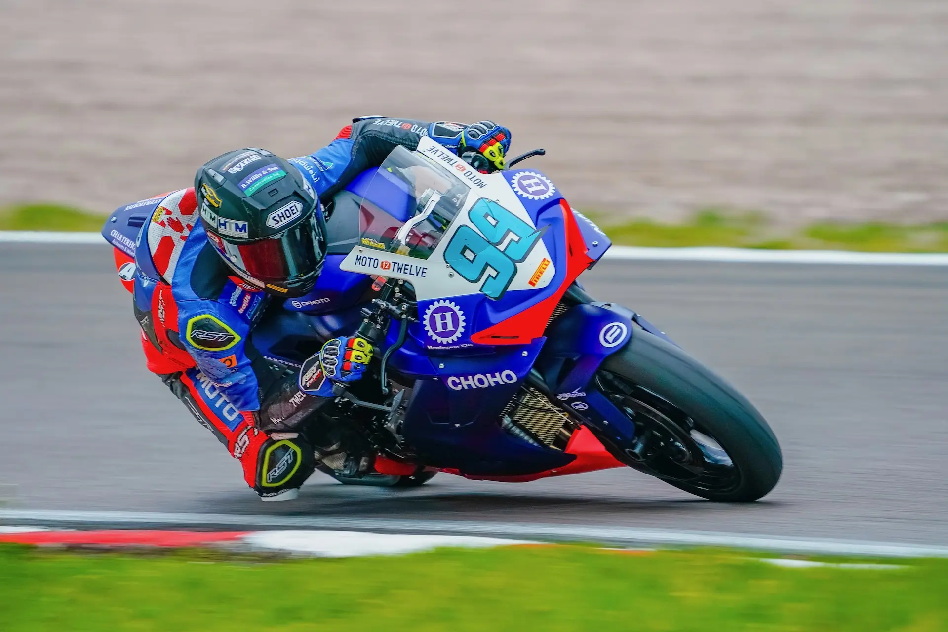 A motorcycle racer in full kit leans sharply into a bend at Oulton Park, riding a blue and red CFMOTO bike with the number 99.