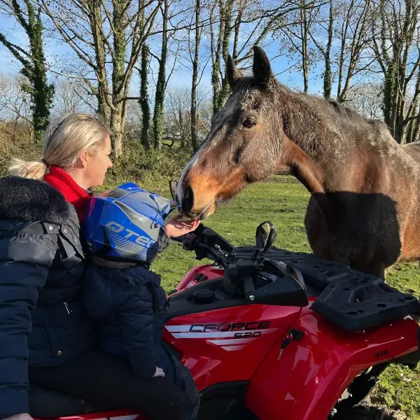 A woman and a child on a red ATV feed a brown horse in a grassy, tree-lined area on a sunny day, enjoying an equestrian moment together.