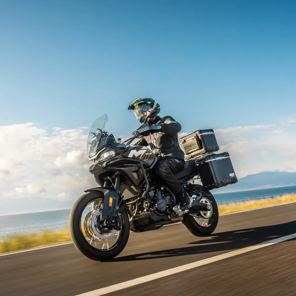 A motorcyclist wearing kit rides a black touring motorbike from a fleet, with panniers, on a coastal road under a clear blue sky.