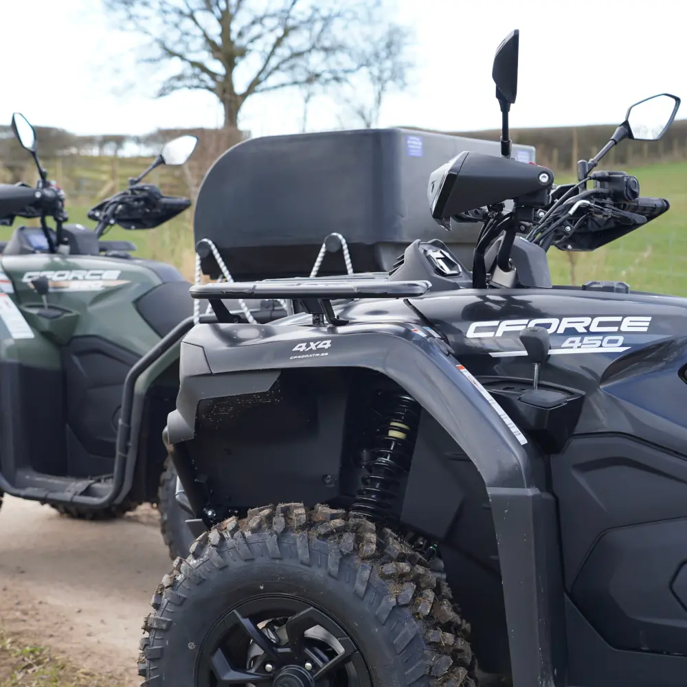 Two CFMOTO CFORCE 450 ATVs from the fleet are parked on a dirt track, with one in the foreground and the other partially visible behind it; a tree and field are in the background.