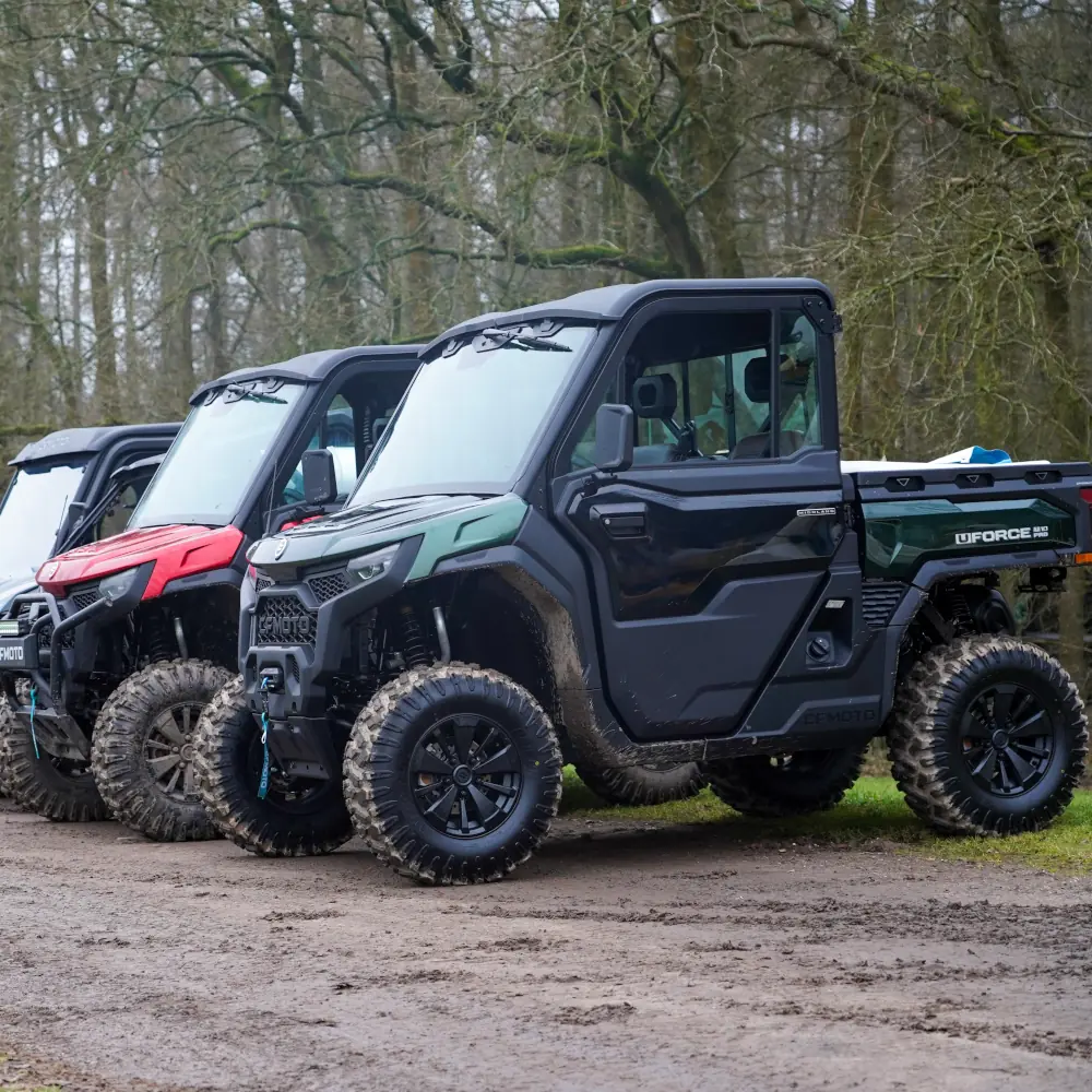 A fleet of three off-road utility vehicles with large, rugged tyres is parked on a dirt path beside a wooded area.