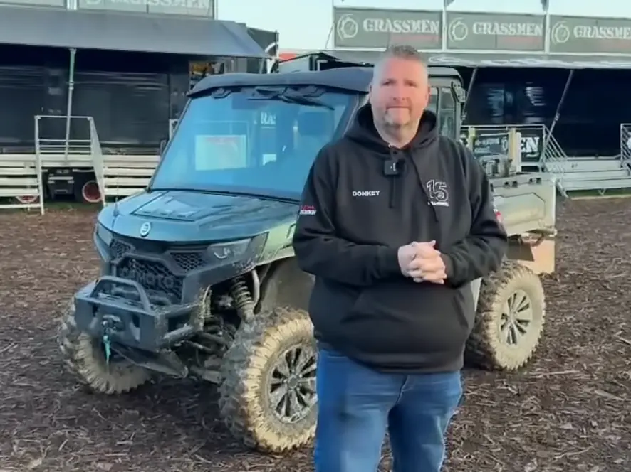 A man wearing a black hoodie stands in front of a muddy CFMOTO CFORCE TECH GEN 4 Concept off-road utility vehicle at an outdoor event with "GRASSMEN" signs in the background.