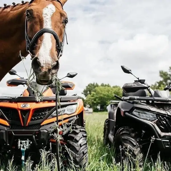 An equestrian horse eating grass stands between two parked all-terrain vehicles in a grassy field under a partly cloudy sky.