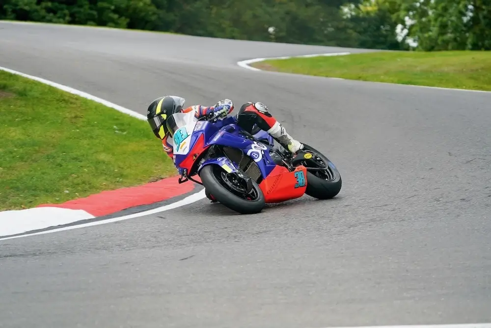 A motorcyclist wearing racing gear, equipped with a CFMOTO T-Box, leans into a sharp turn on a racetrack, surrounded by grass and trees.