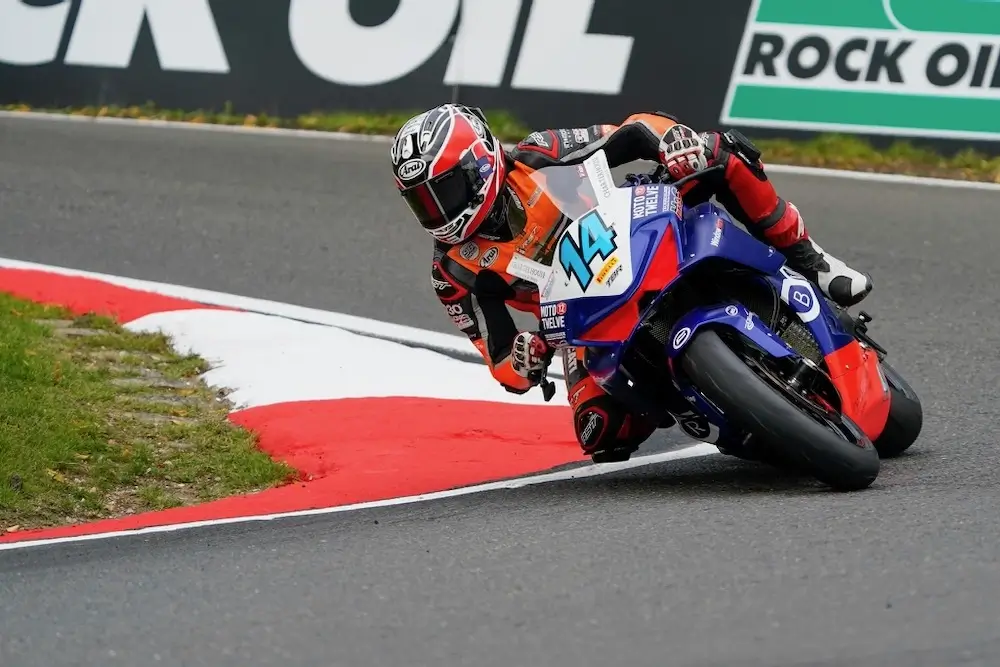 Motorcycle racer in red and black gear leans sharply into a turn on a racetrack, riding a blue and red CFMOTO T-Box bike numbered 14. Sponsor banners are visible in the background.
