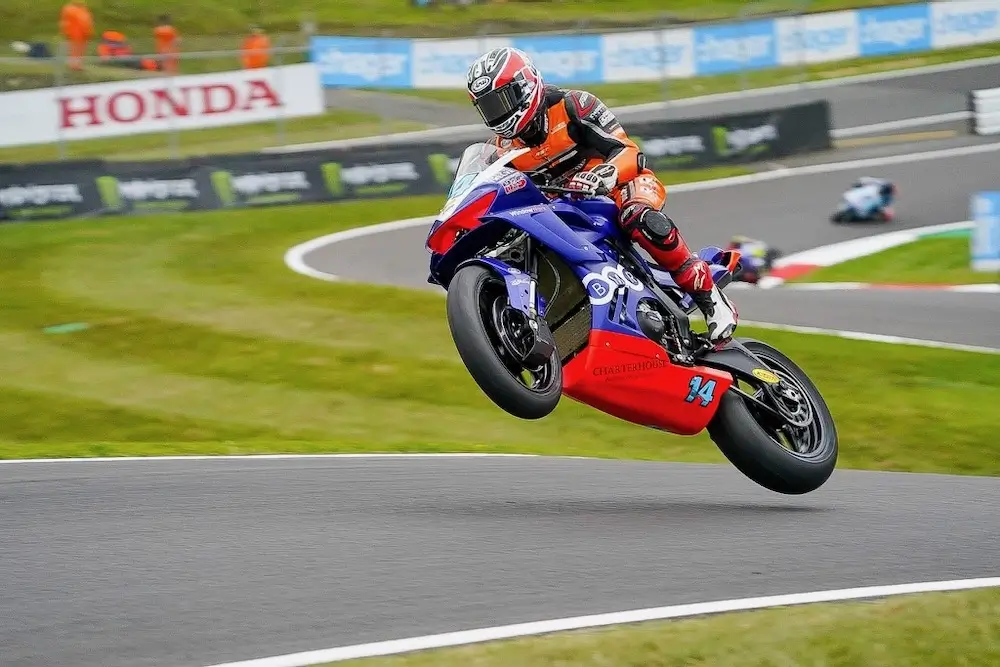 A motorcycle racer in full gear rides a blue and red bike with the number 34, likely equipped with a CFMOTO T-Box, lifting the front wheel off the ground on a racetrack as other riders and track marshals look on in the background.