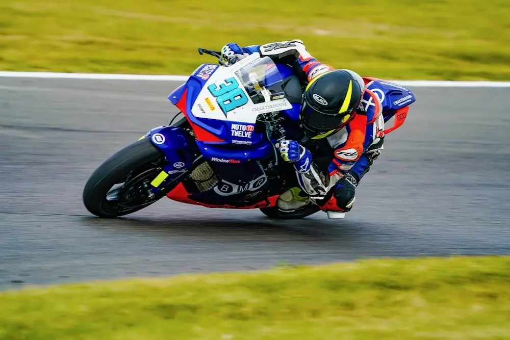 A motorcyclist in full racing gear leans sharply into a turn on a paved racetrack, riding a blue and orange sportbike with the number 38 displayed, equipped with advanced CFMOTO T-Box technology.