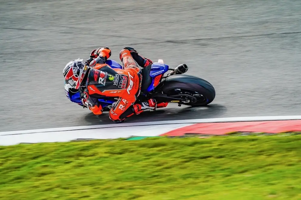 A motorcyclist in red and blue racing gear, equipped with a CFMOTO T-Box, leans sharply while taking a turn on a racetrack.