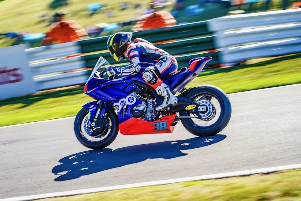 A motorcycle racer in blue and white gear rides a blue sportbike equipped with a CFMOTO T-Box at high speed on a racetrack, with a blurred background and spectators in the distance.