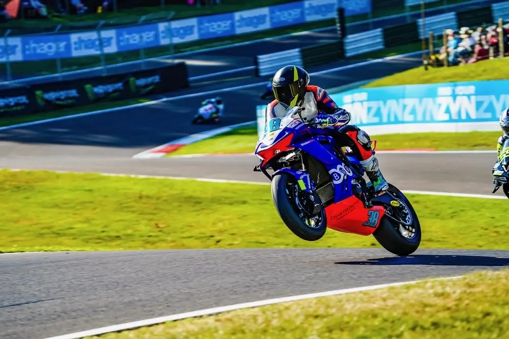 A motorcycle racer equipped with a CFMOTO T-Box lifts the front wheel off the ground while speeding around a bend on a racetrack, with another rider visible in the background.