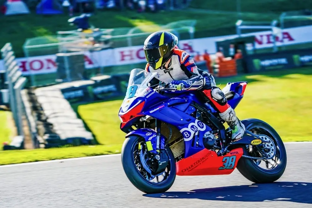 A motorcyclist in racing gear rides a blue and red sport bike, featuring CFMOTO T-Box technology, on a racetrack with blurred Honda banners and green grass in the background.