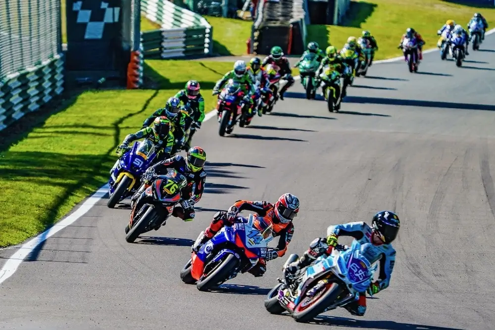 A group of motorcycle racers, some equipped with CFMOTO T-Box technology, speed around a bend on a paved track during a race, with green grass and barriers along the sides.