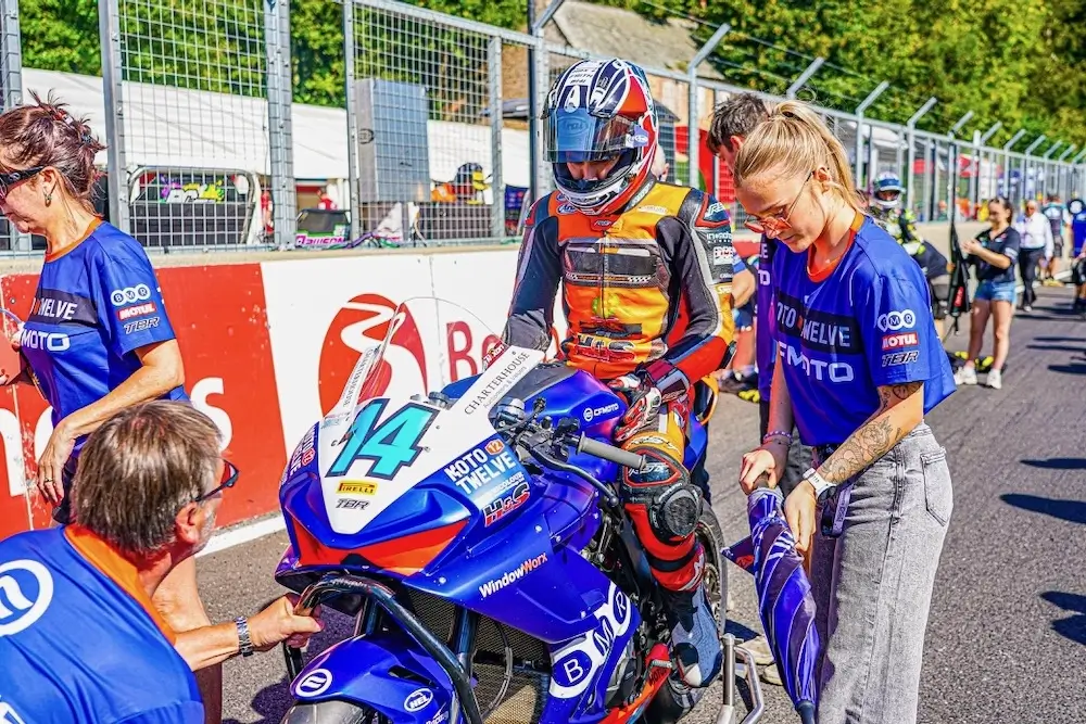 A motorcycle racer in full gear sits on a blue CFMOTO T-Box race bike numbered 14, surrounded by team members preparing on a racetrack.