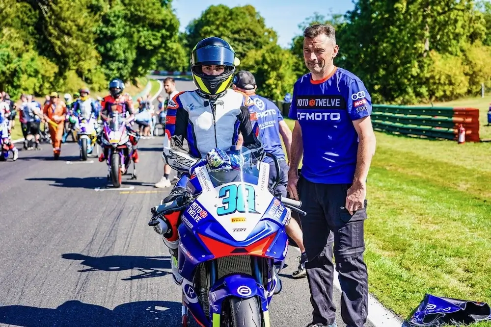 Motorcycle racer in full gear sits on bike number 31 equipped with a CFMOTO T-Box at a racetrack, with a team member beside him; other racers are lined up in the background.