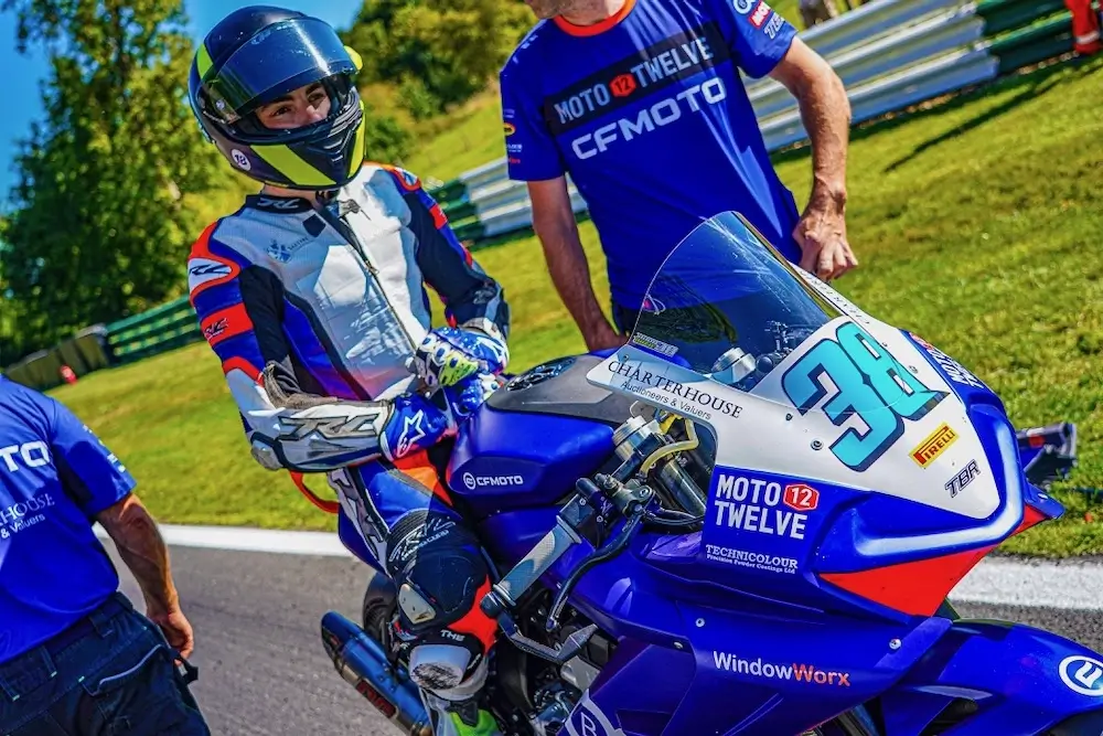 A motorcycle racer wearing full gear sits on a blue CFMOTO T-Box racing bike numbered 38, flanked by two team members in blue shirts, on a sunny outdoor track.