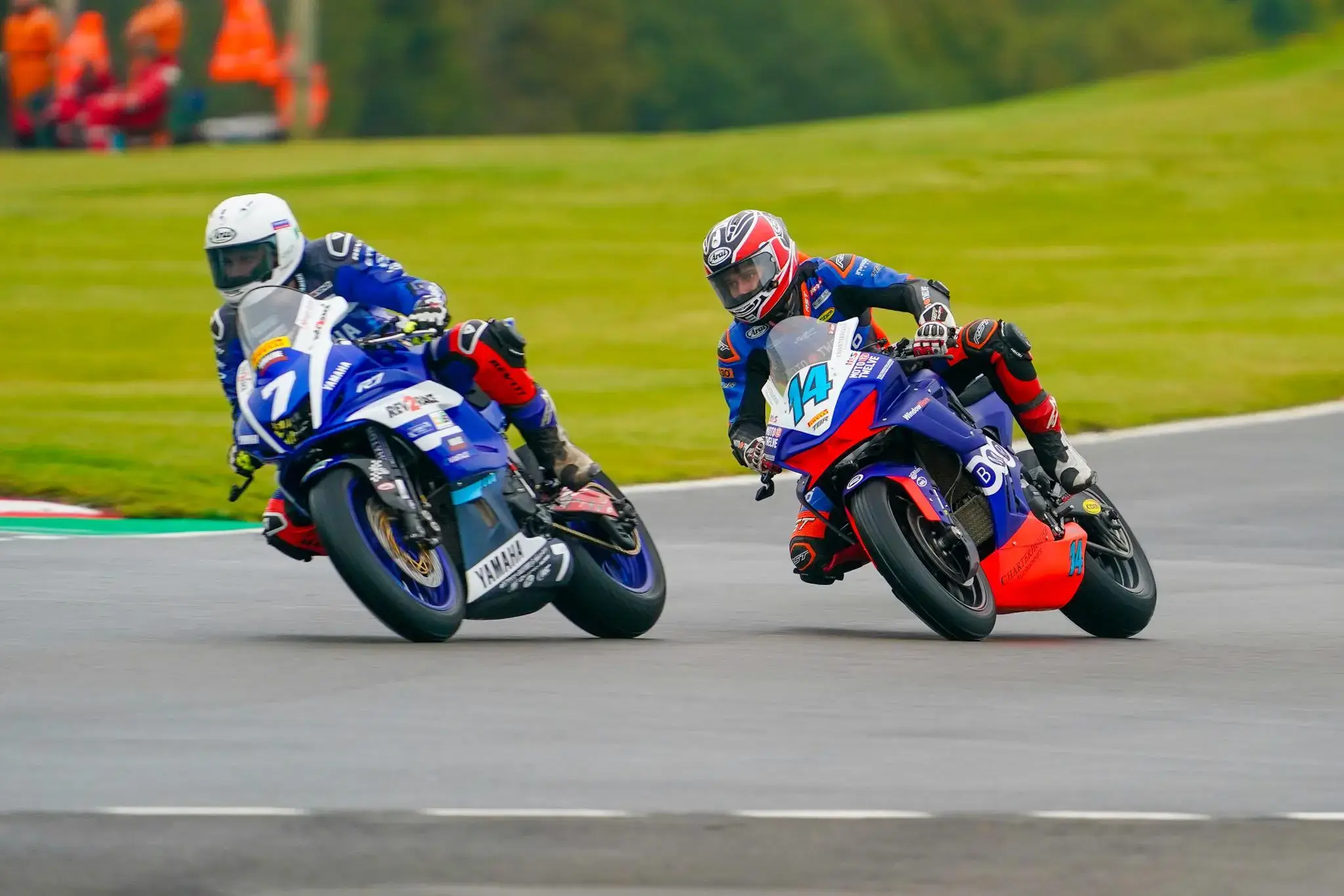Two motorcyclists in racing gear lean into a turn on the racetrack during the National Sportbike Championship, with grass and safety crew visible in the background.
