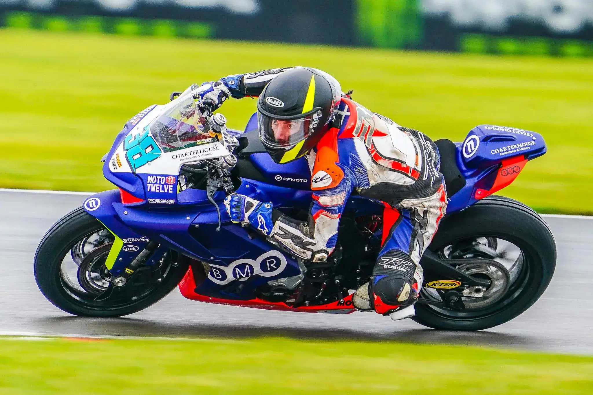 A motorcycle racer in full gear leans into a turn on a blue racing bike during the National Sportbike Championship, with green grass lining the track in the background.