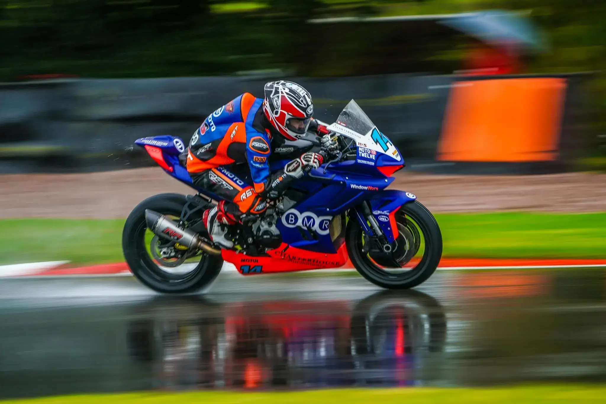 A motorcyclist in racing gear rides a blue and orange motorcycle at high speed on a wet race track during the R9 Race, with a blurred background indicating motion.