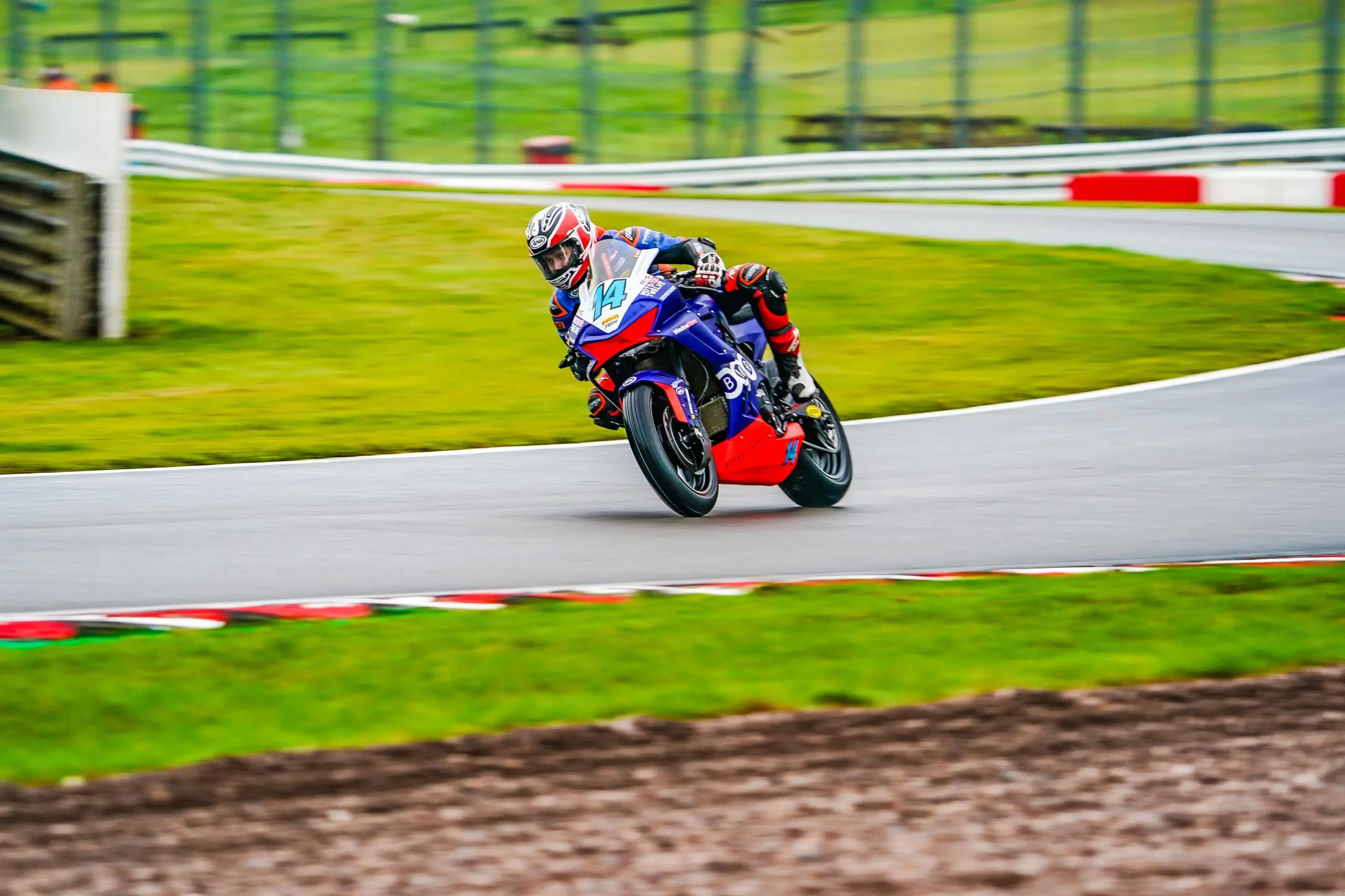 A motorcyclist in racing gear rides a sportbike around a curved track during the National Sportbike Championship, leaning into the turn on a paved road surrounded by grass.