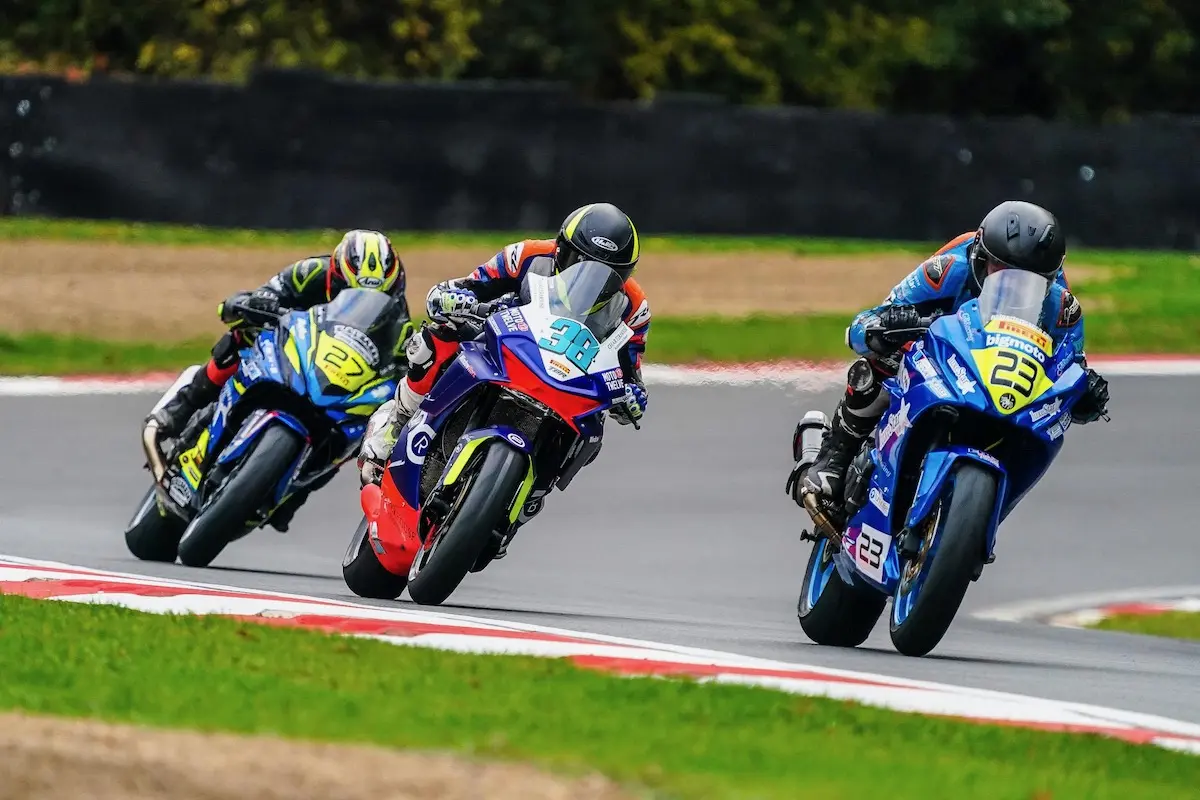 Three motorcycle racers on sportbikes lean into a turn on a racetrack, wearing full racing gear and helmets, with green grass and trees in the background—capturing the thrill of Moto Twelve.