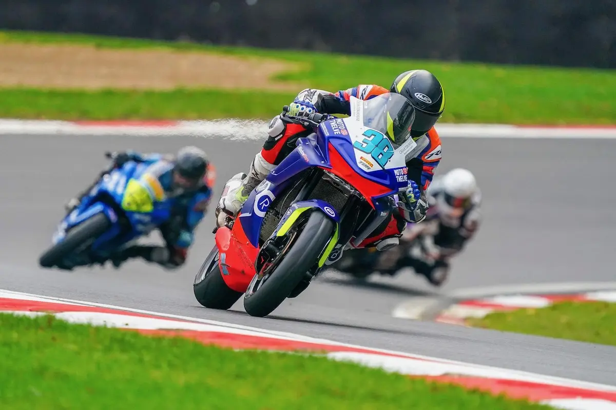 A CFMOTO motorcycle racer leans into a curve on the track, followed closely by two other riders, aiming for the National Sportbike Podium, with grass and asphalt visible in the background.