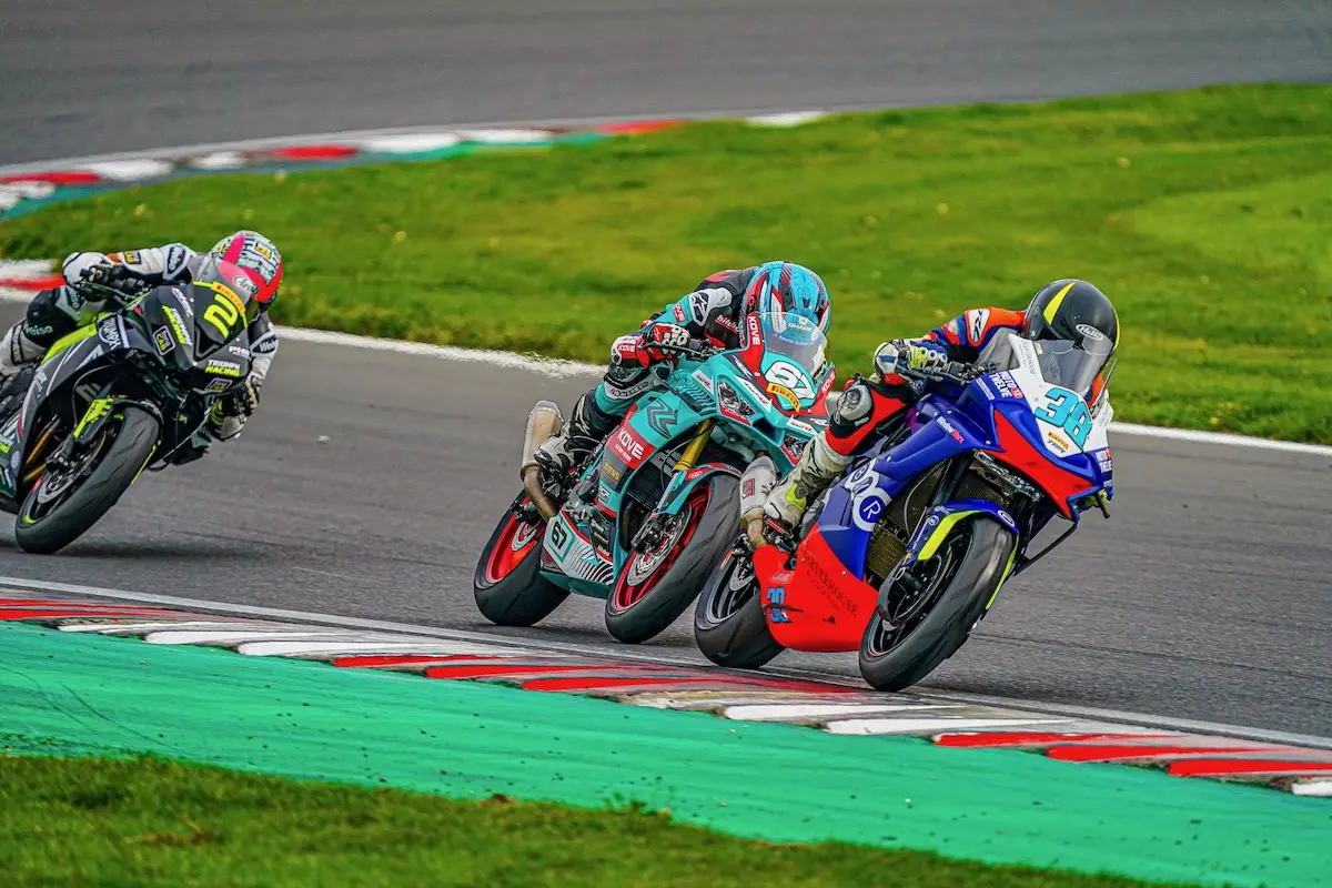 Three motorcycle racers lean into a curve on a racetrack, closely competing in colorful racing suits and CFMOTO helmets, each determined to claim a National Sportbike Podium spot.
