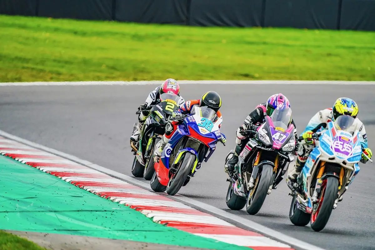 Four motorcyclists race closely on a paved track, leaning into a curve during a Moto Twelve event, with a grassy area and barriers in the background.
