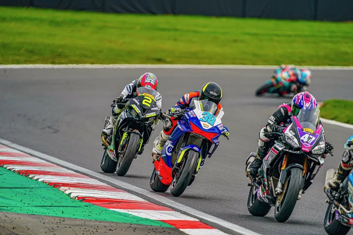 Three CFMOTO motorcycle racers take a sharp turn on a racetrack, competing fiercely for the Sportbike Podium as another racer lags behind in the background.