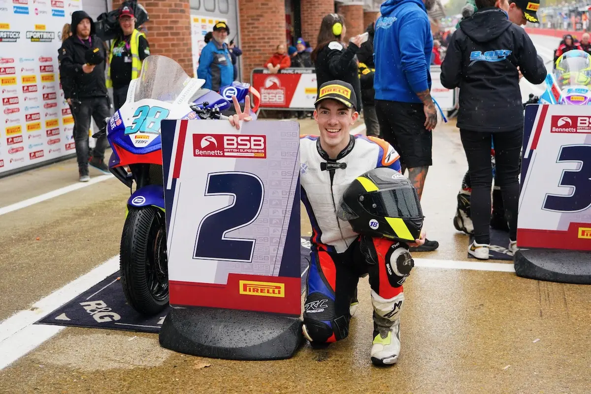 A motorcycle racer in racing gear kneels next to a “2” National Sportbike Podium sign, holding a helmet; his CFMOTO bike and other people are visible in the wet pit lane background.