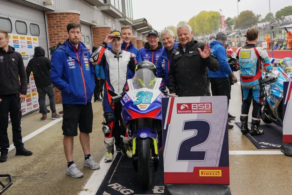 IMG-20251019-WA0091 A group of people stand behind a CFMOTO motorcycle marked with the number 38, next to a sign displaying "2" at a Moto Twelve racing event.