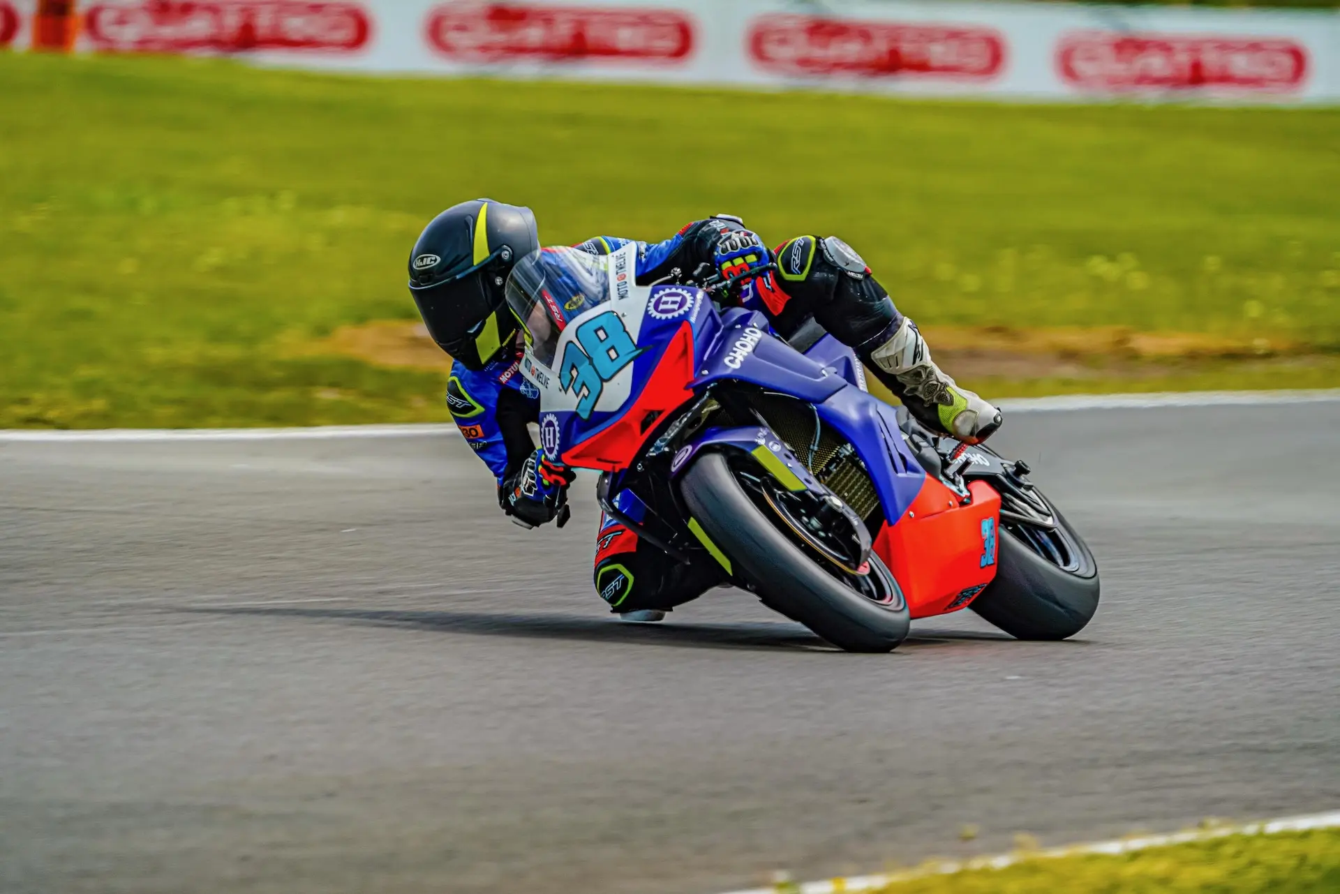 A motorcycle racer in a blue and black suit leans into a bend at Oulton Park, with blurred grass and advertisements in the background, showcasing the thrill of Moto Twelve racing.