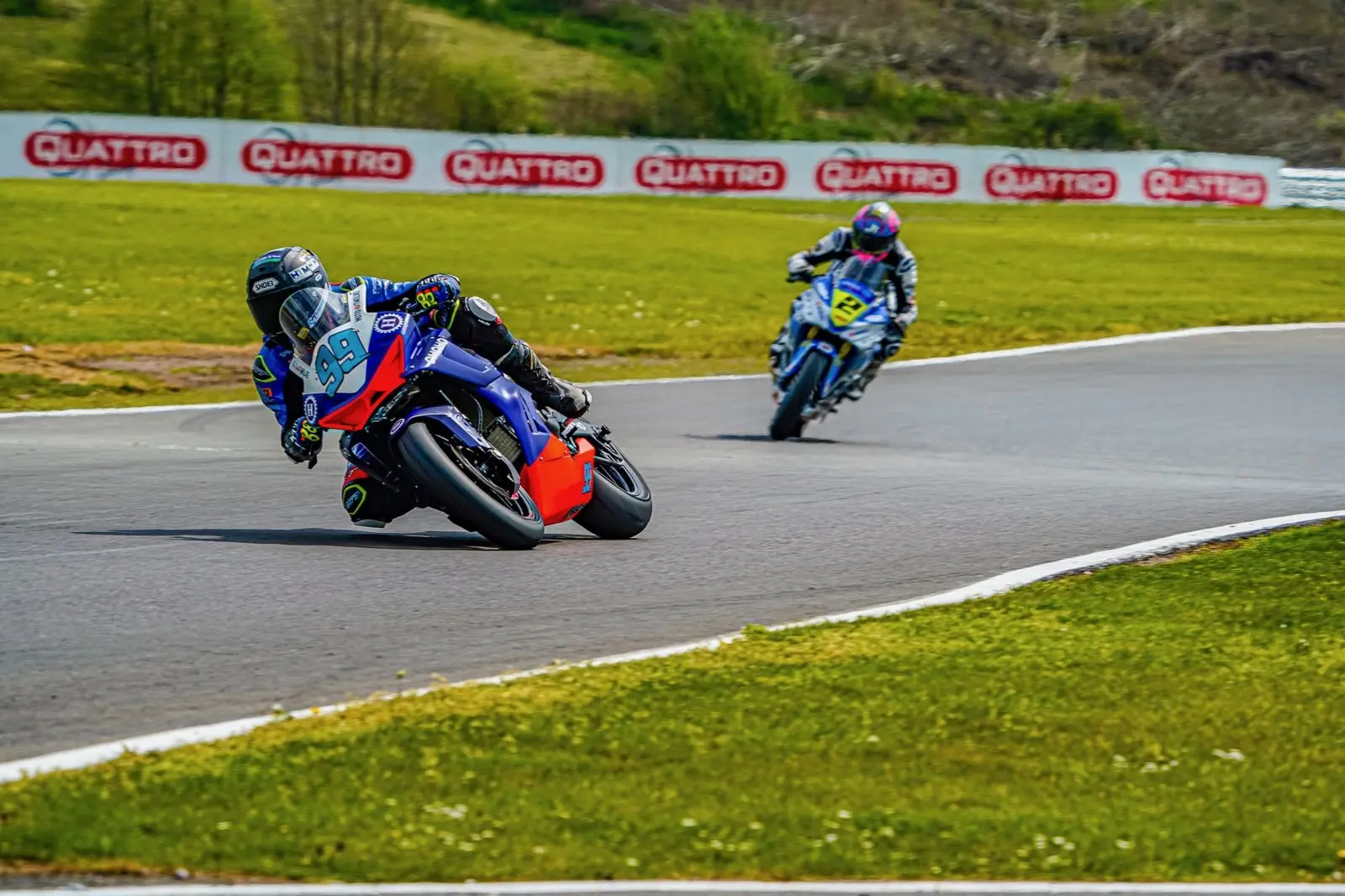 Two motorcycle racers in blue suits lean into a bend at Oulton Park, the iconic circuit with grassy surroundings and advertising hoardings, competing on their CFMOTO bikes.