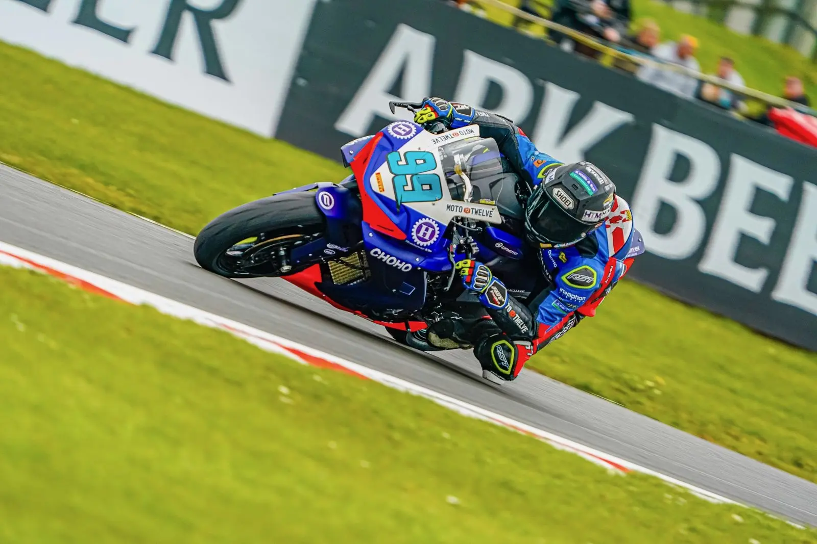 A motorcycle racer in blue kit leans into a bend at Oulton Park on a racetrack, surrounded by green grass and sponsor banners, showcasing the thrill of Moto Twelve and CFMOTO excellence.