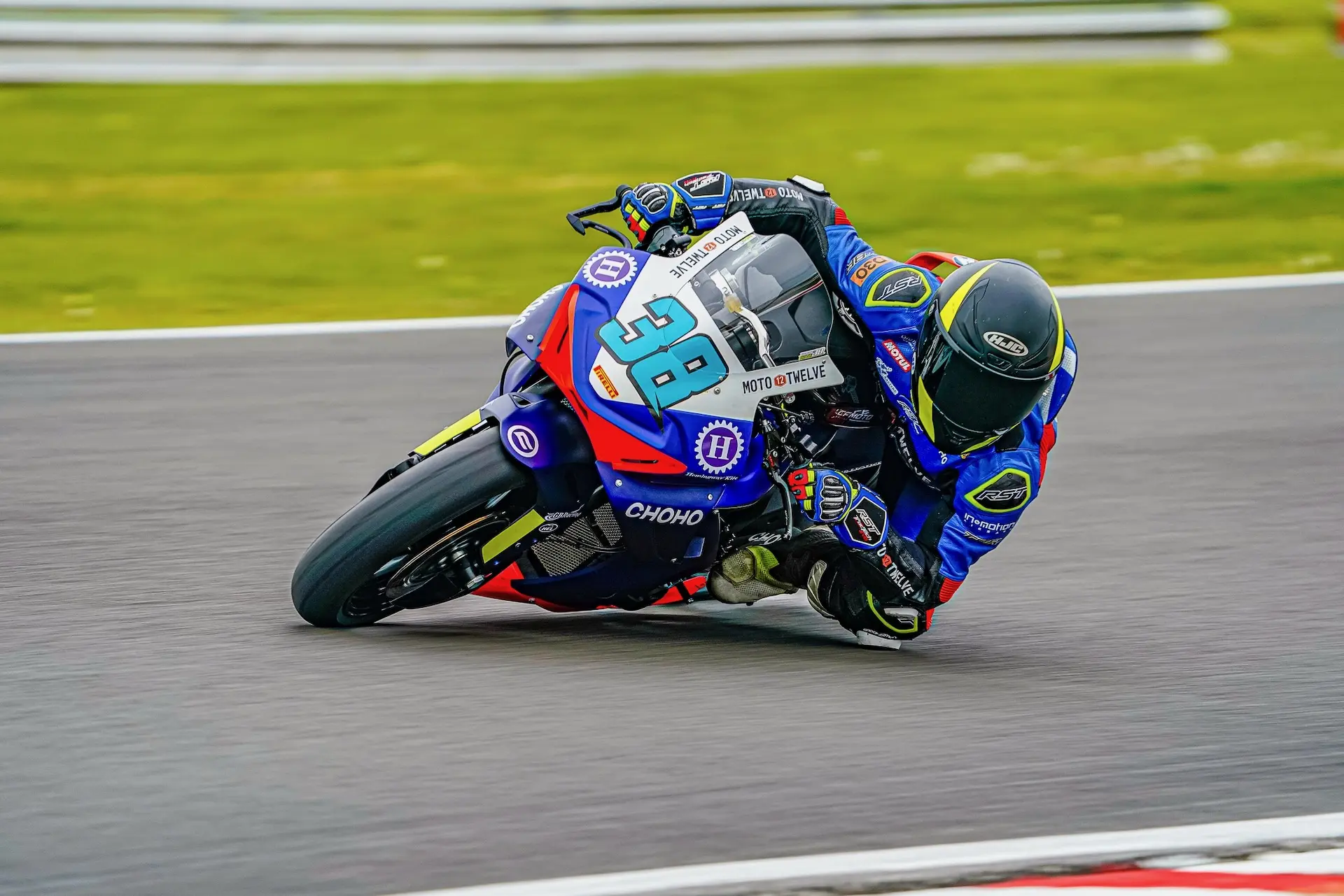 A motorcycle racer in blue and red kit leans sharply on the Oulton Park circuit, riding a CFMOTO bike marked with the number 38.