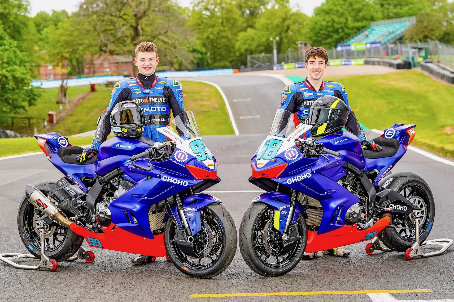 Two male motorcycle racers in racing suits stand next to blue and red CFMOTO race bikes on a track at Oulton Park, smiling at the camera with greenery and stands in the background.