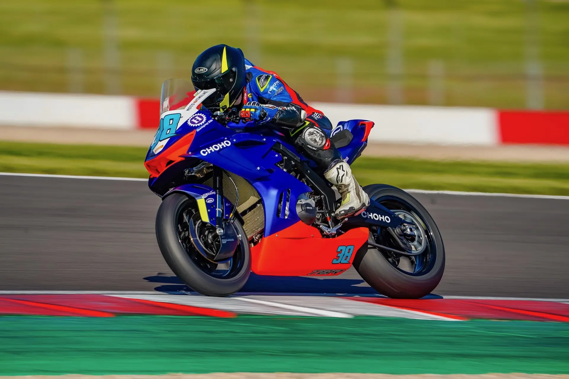 A motorcycle racer in full gear rides a blue and red CFMOTO sports bike, leaning into a turn at Donington Park with a blurred green and red background.