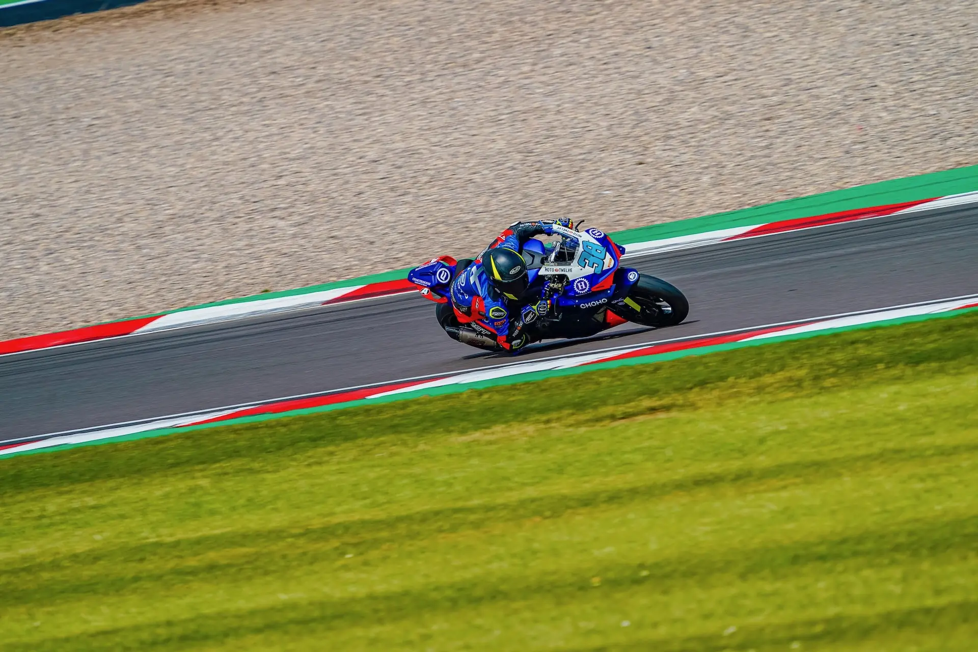 A motorcycle racer in blue gear leans into a turn on the Donington Park racetrack, surrounded by grass and bordered by red, white, and green kerbs.