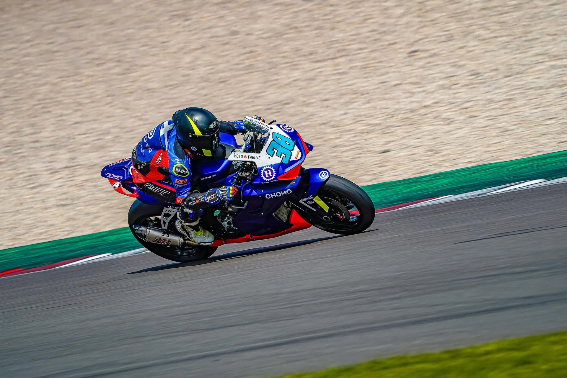 A motorcycle racer in blue and red gear leans into a turn on the Donington Park racetrack, with a gravel run-off area in the background.