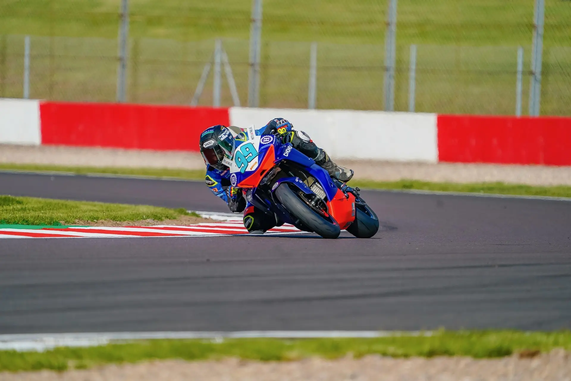 A Moto Twelve motorcyclist leans into a bend on the Donington Park racetrack, with green grass and a red-and-white barrier in the background.
