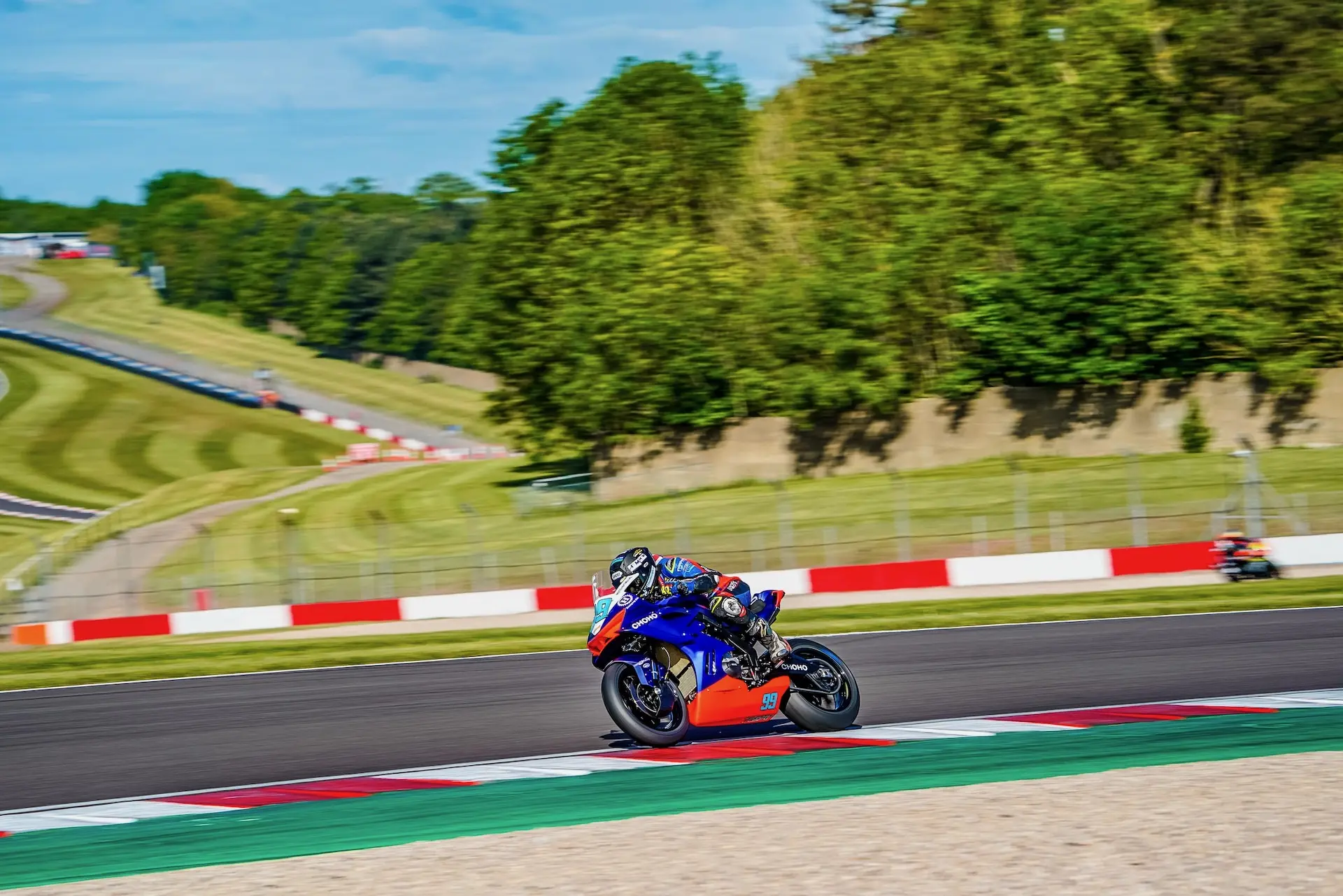 A motorcyclist in racing gear rides a blue and red CFMOTO sports motorbike on a racetrack at Donington Park, with green grass and trees in the background.