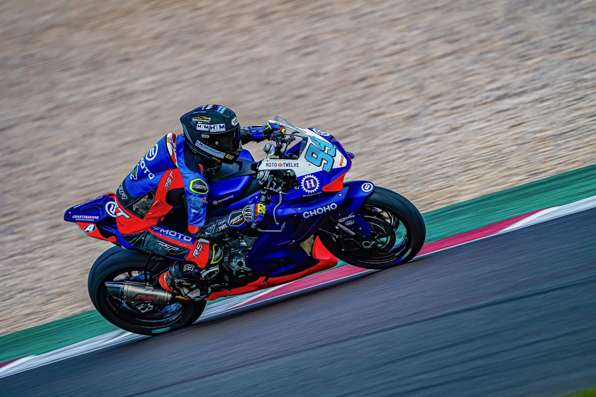 A motorcycle racer in blue and red kit rides a CFMOTO sport bike at high speed on the Donington Park racetrack.