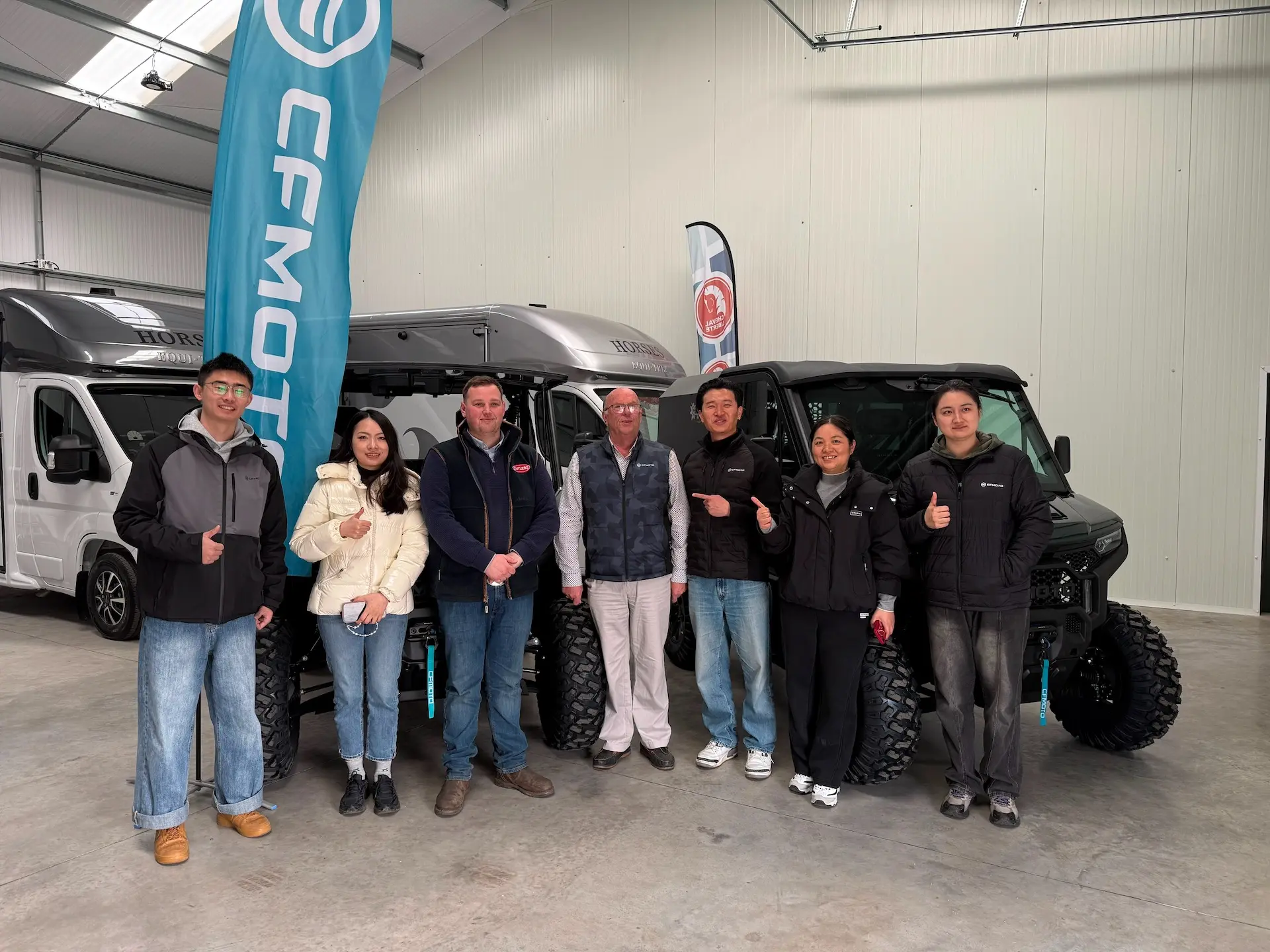 A group of seven people stand indoors in front of off-road vehicles, posing for a photo with a CFMOTO flag visible on the left—highlighting their connection to agricultural UTVs.