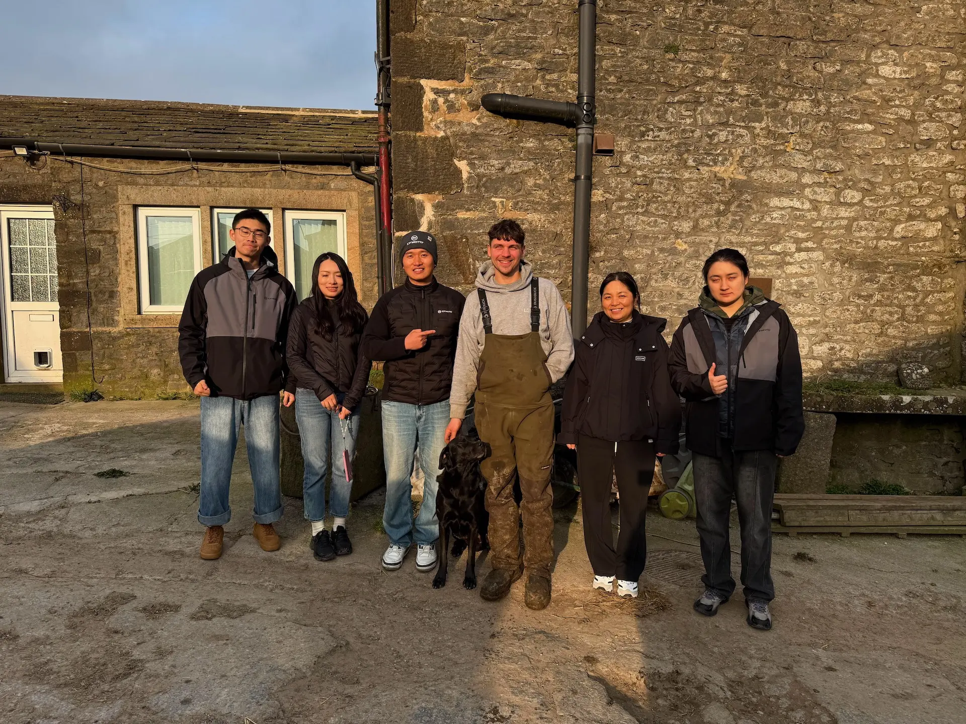 Six people and a black dog stand outdoors in front of a stone building, posing for a group photo in casual clothing and jackets—typical of UK farmers gathering to discuss agricultural UTVs.