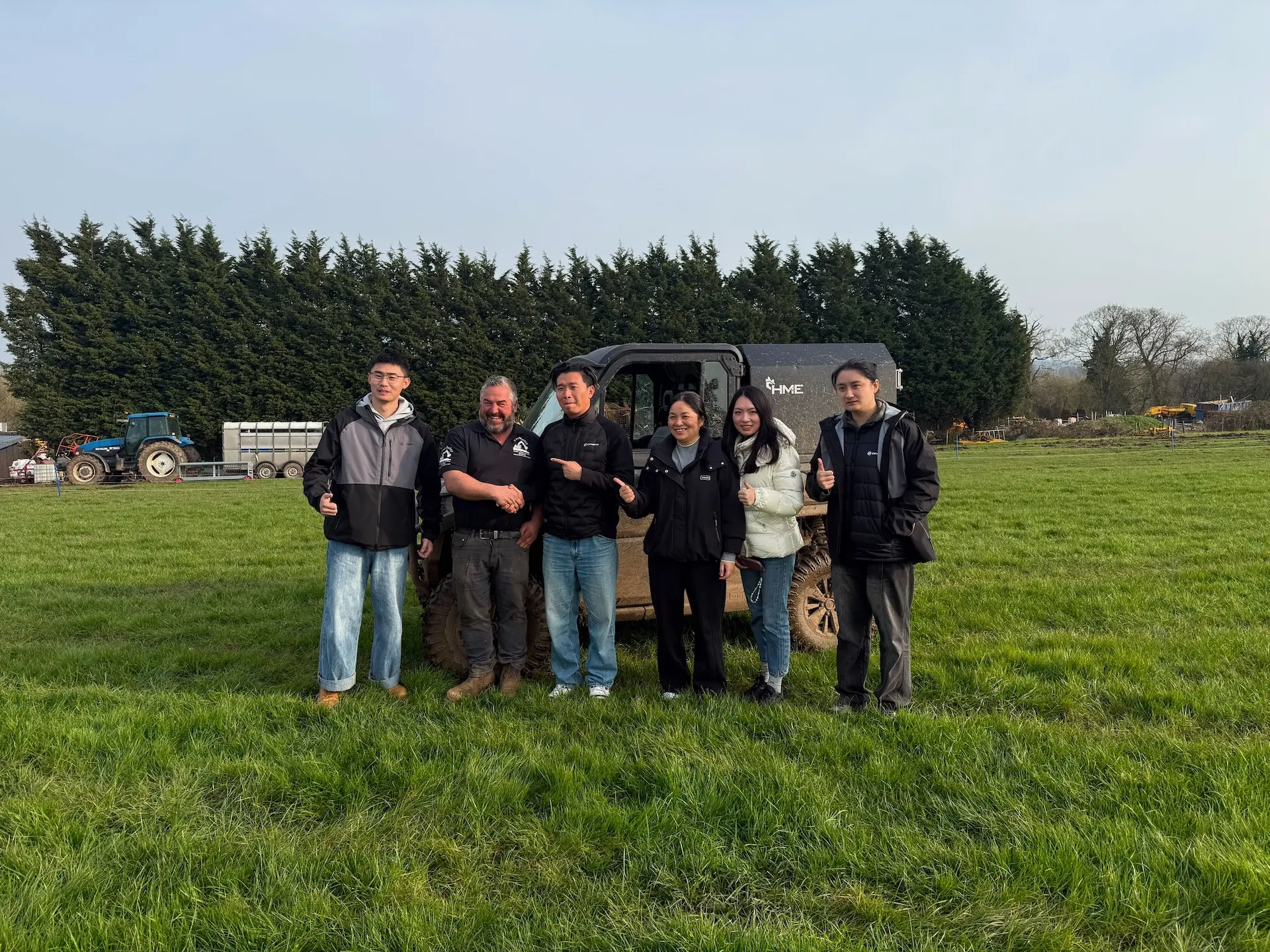 Six people stand in a row on grass, posing for a photo with thumbs up; a CFMOTO agricultural UTV and trees are visible in the background on a clear day.
