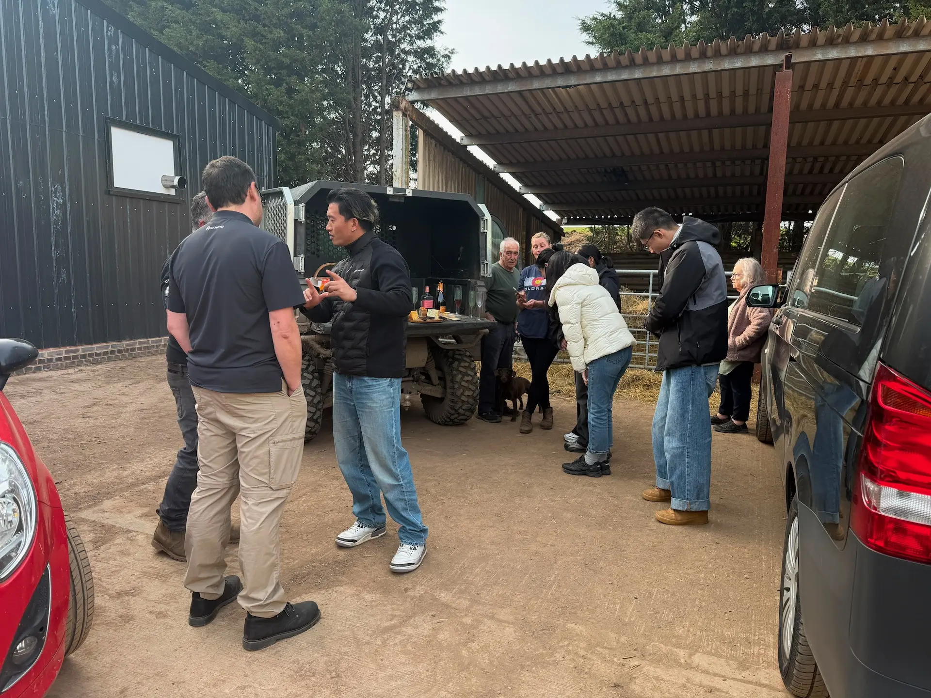 A group of people, including UK farmers, stand and chat near parked cars, agricultural UTVs, and a table with refreshments outside a metal building.